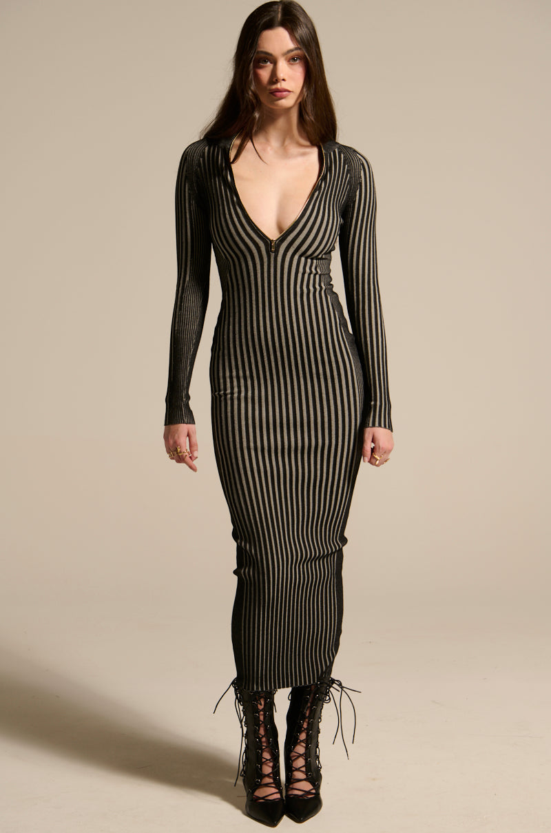 A woman with long brown hair faces forward, wearing the TAKE ME HOME KNIT MAXI DRESS, which has ribbed texture and black and beige vertical stripes with a deep V-neckline. She pairs it with black lace-up high-heeled shoes against a plain background.