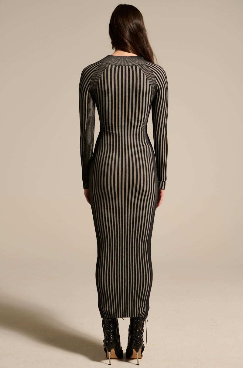 A woman faces away wearing the TAKE ME HOME KNIT MAXI DRESS, a fitted ribbed maxi with vertical black and beige stripes, paired with black lace-up heeled boots. Her long dark hair is loose against a plain beige background.