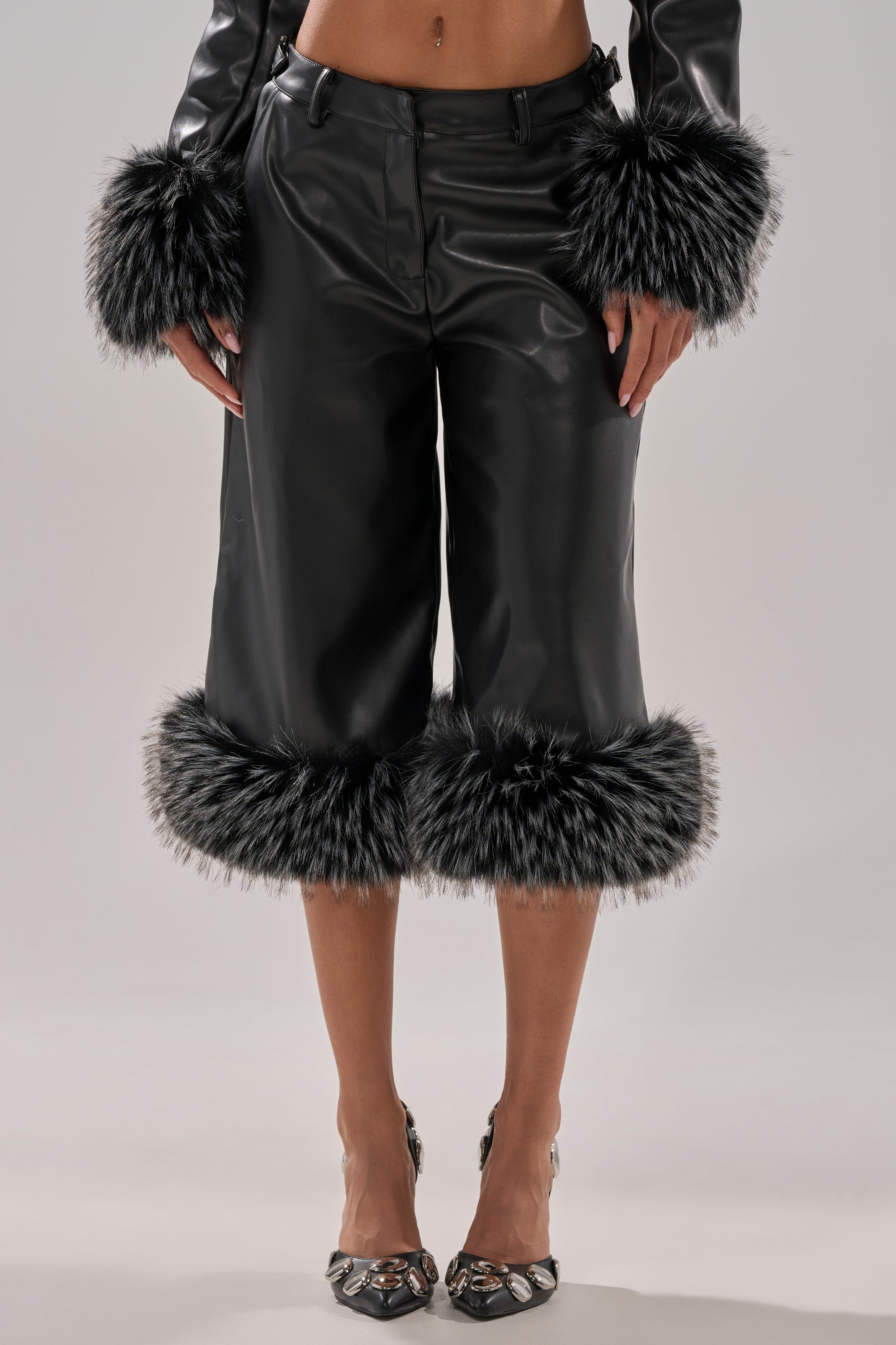 A person wears the MIND YOUR OWN FUR TRIM BERMUDA SHORTS, featuring a tailored waistband and black-and-white faux fur trim, paired with a matching faux fur cuffed jacket and pointed metallic shoes.