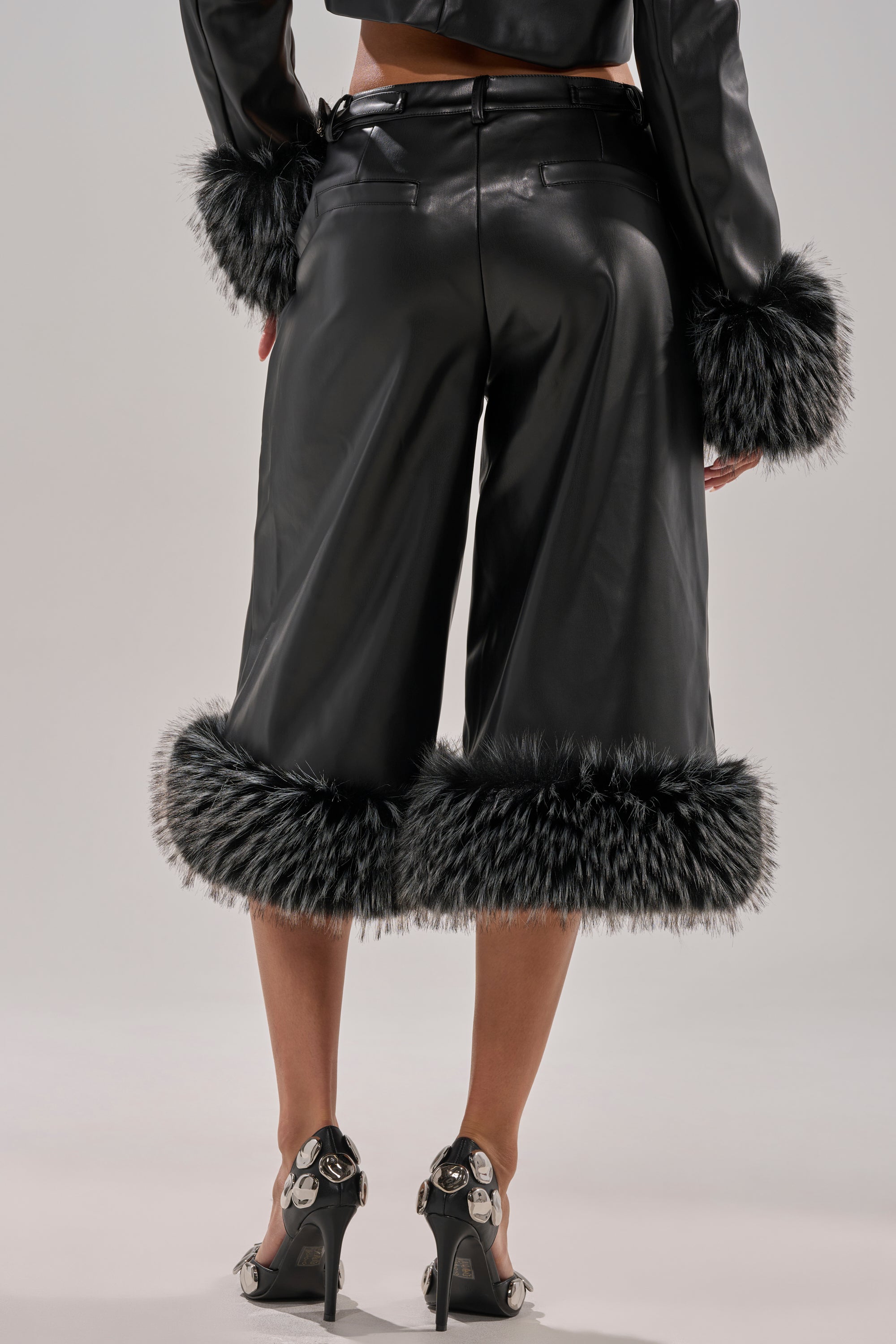 Someone stands against a light background wearing MIND YOUR OWN FUR TRIM BERMUDA SHORTS with black and white faux fur trim and matching cuffs, paired with black heeled sandals adorned with metallic skull decorations.