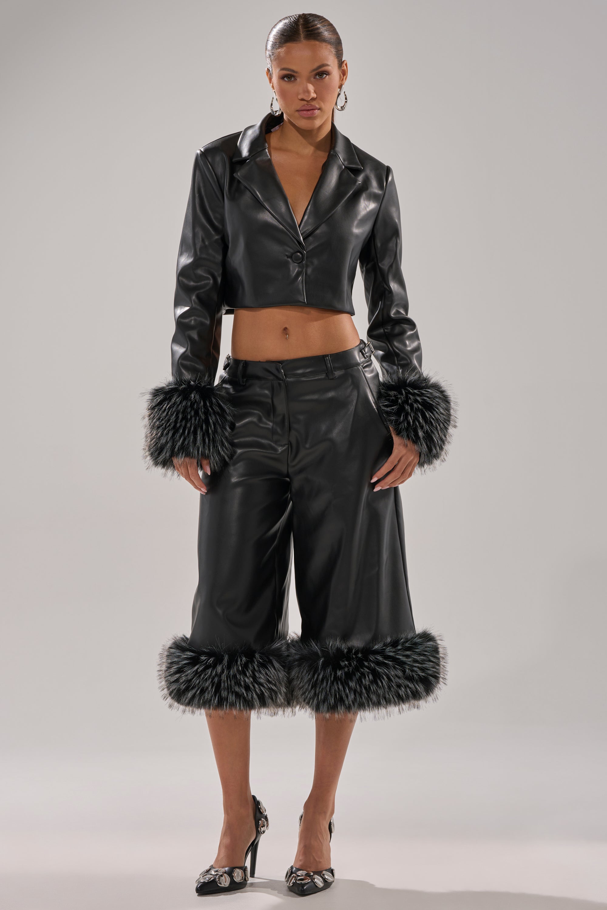 A woman wears MIND YOUR OWN FUR TRIM BERMUDA SHORTS and a matching faux leather cropped jacket, both with black-and-white fur trim. She completes the outfit with hoop earrings and coordinating black-and-white heels.
