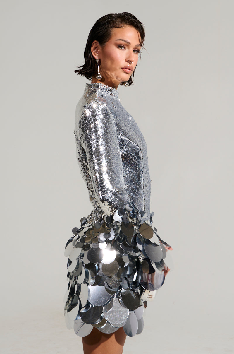 A person wearing THE MOMENT SEQUIN MINI DRESS, featuring reflective sequin detailing in shiny silver, stands in profile against a plain background. They have short, slicked-back hair and statement earrings, holding a small clutch.