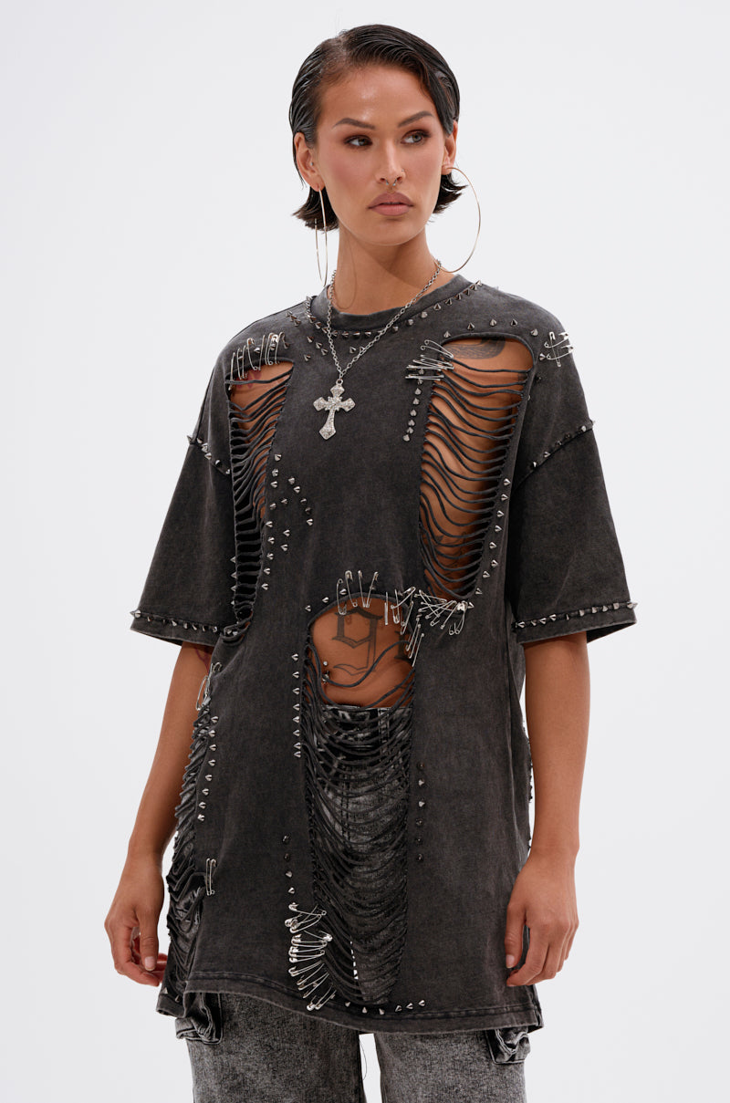 A person with wet-look slicked-back hair wears the MARGOT DISTRESSED STUDDED SHORT SLEEVE TEE, featuring large slashes, safety pins, and chains over a striped top, accessorized with hoop earrings and a cross necklace.