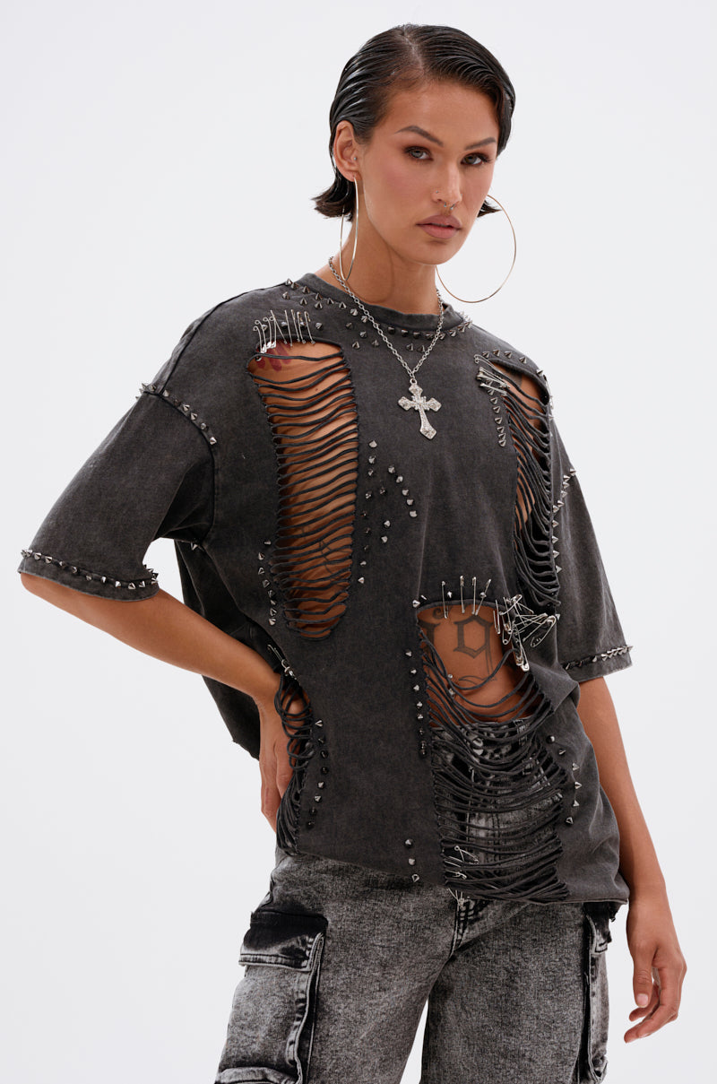 Against a plain background, an individual with slicked-back hair models the MARGOT DISTRESSED STUDDED SHORT SLEEVE TEE—featuring rips and mesh details—paired with large hoop earrings, a cross necklace, and faded worn jeans.