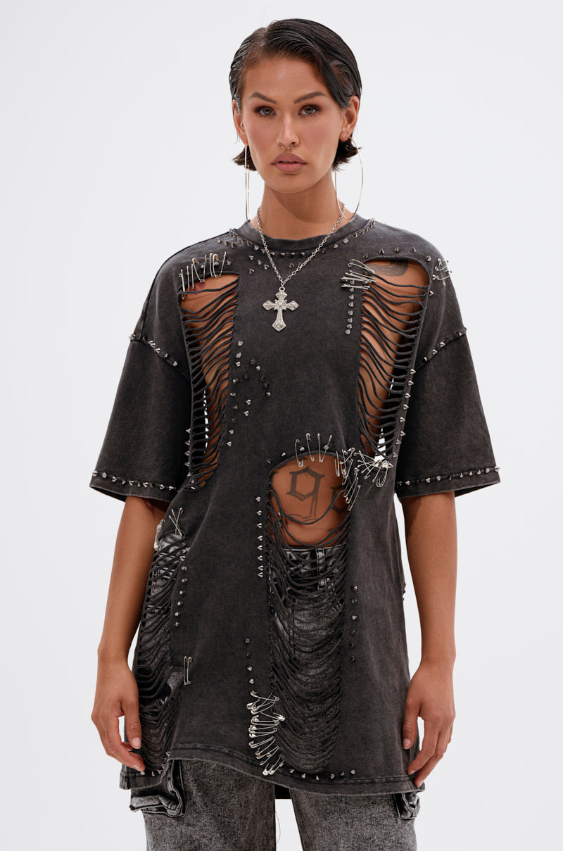 A person wearing the MARGOT DISTRESSED STUDDED SHORT SLEEVE TEE with large rips, safety pins, chains, and metal accents pairs it with hoop earrings and a cross necklace, standing against a plain white background.