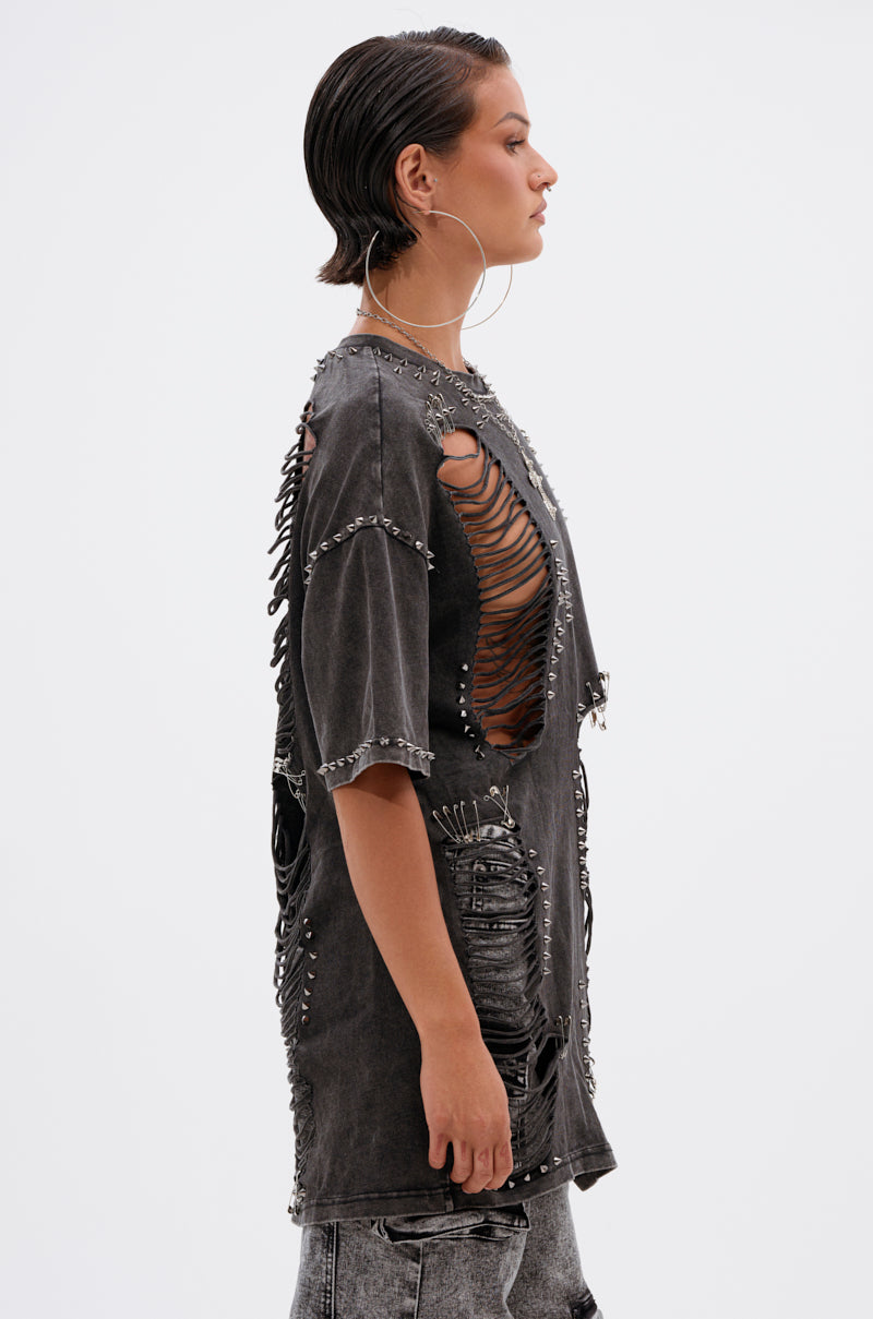 A person with slicked-back hair and large hoop earrings wears the MARGOT DISTRESSED STUDDED SHORT SLEEVE TEE, featuring rips, metal studs, and cutouts, paired with light-wash distressed jeans. The background is plain white.