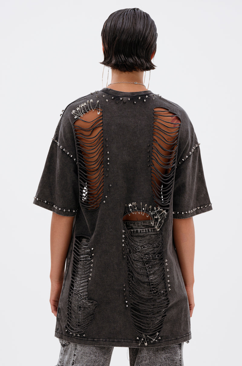 A person is seen from behind wearing the MARGOT DISTRESSED STUDDED SHORT SLEEVE TEE, featuring large shredded holes and safety pins, styled with gray pants. Their hair is short and wet-look, set against a plain background.