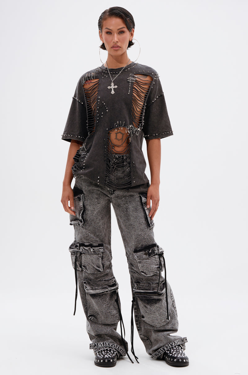 Wearing the MARGOT DISTRESSED STUDDED SHORT SLEEVE TEE, grey cargo pants, studded shoes, large hoop earrings, and a cross necklace, a person poses against a plain white background.