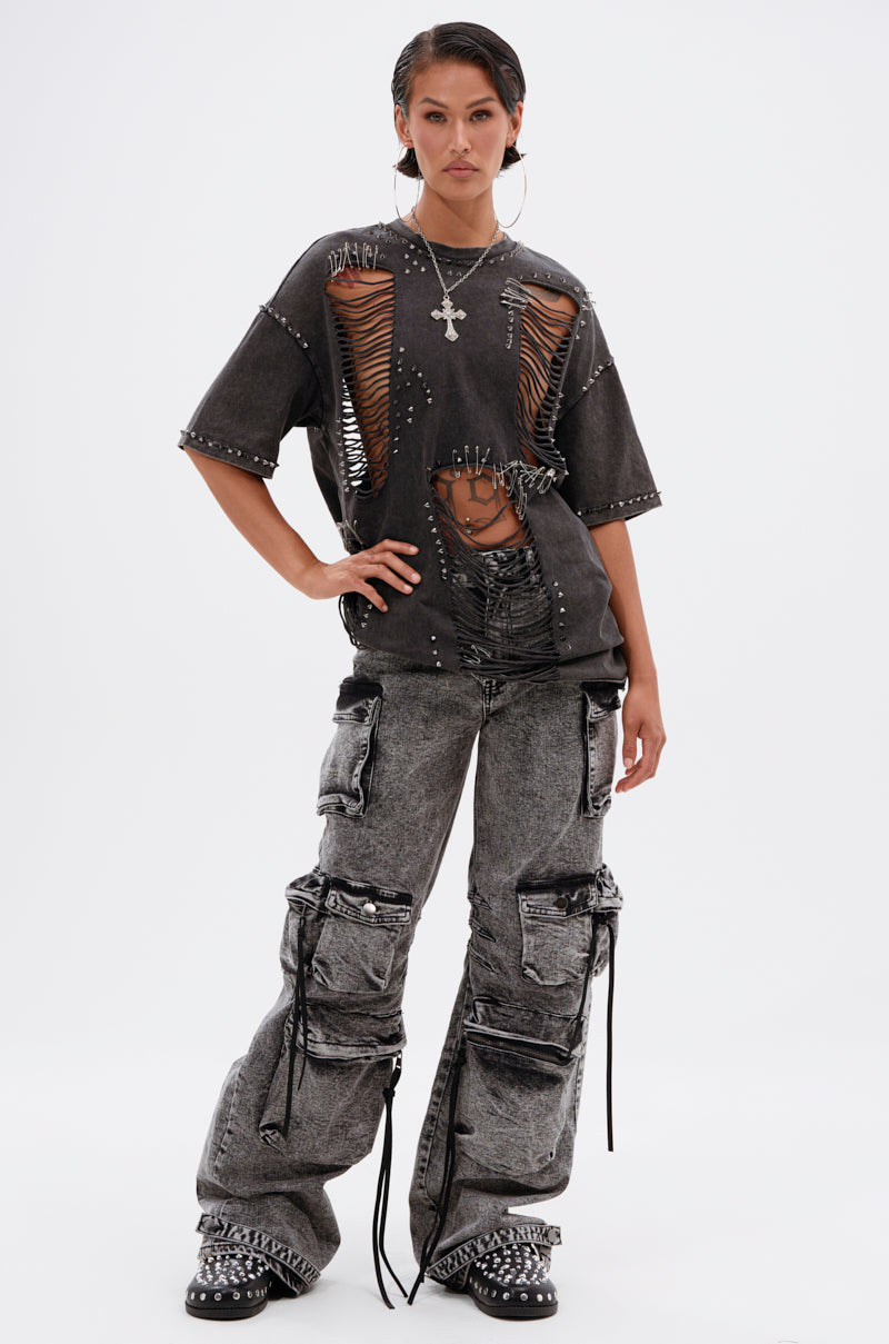 A person stands confidently in the MARGOT DISTRESSED STUDDED SHORT SLEEVE TEE with large rips, paired with gray cargo pants, chunky shoes, hoop earrings, and a cross necklace against a plain white background.