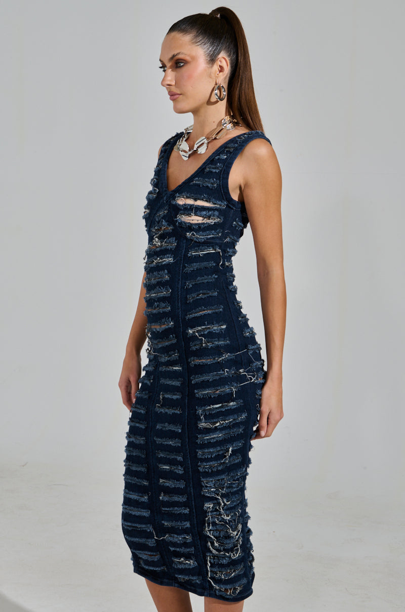 A person wearing the "She’s Got It Shredded Denim Midi Dress in Dark Blue" poses against a plain background, styled with a high ponytail and statement jewelry including large earrings and a chunky necklace.