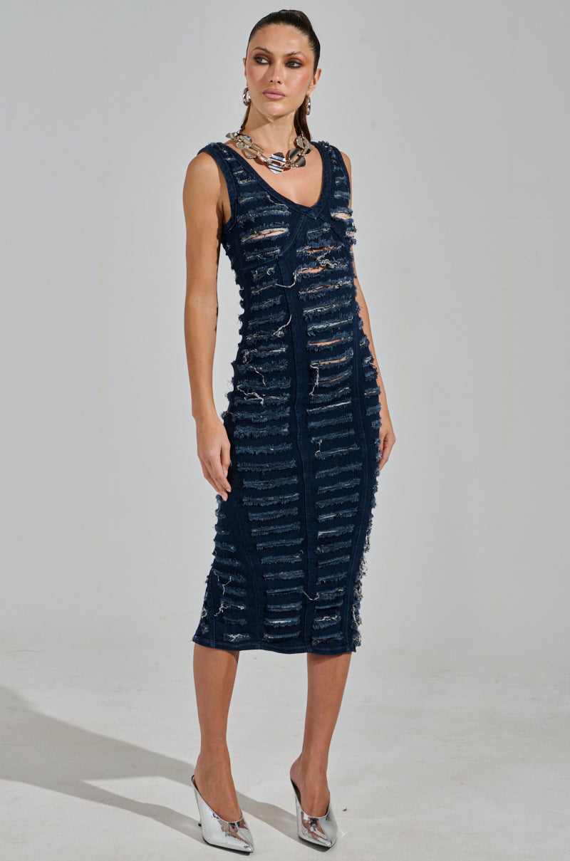 A person poses in the SHE'S GOT IT SHREDDED DENIM MIDI DRESS IN DARK BLUE with metallic heels and a chunky necklace. Their hair is tied back, and the neutral background highlights the sleeveless, knee-length, curve-hugging dress.