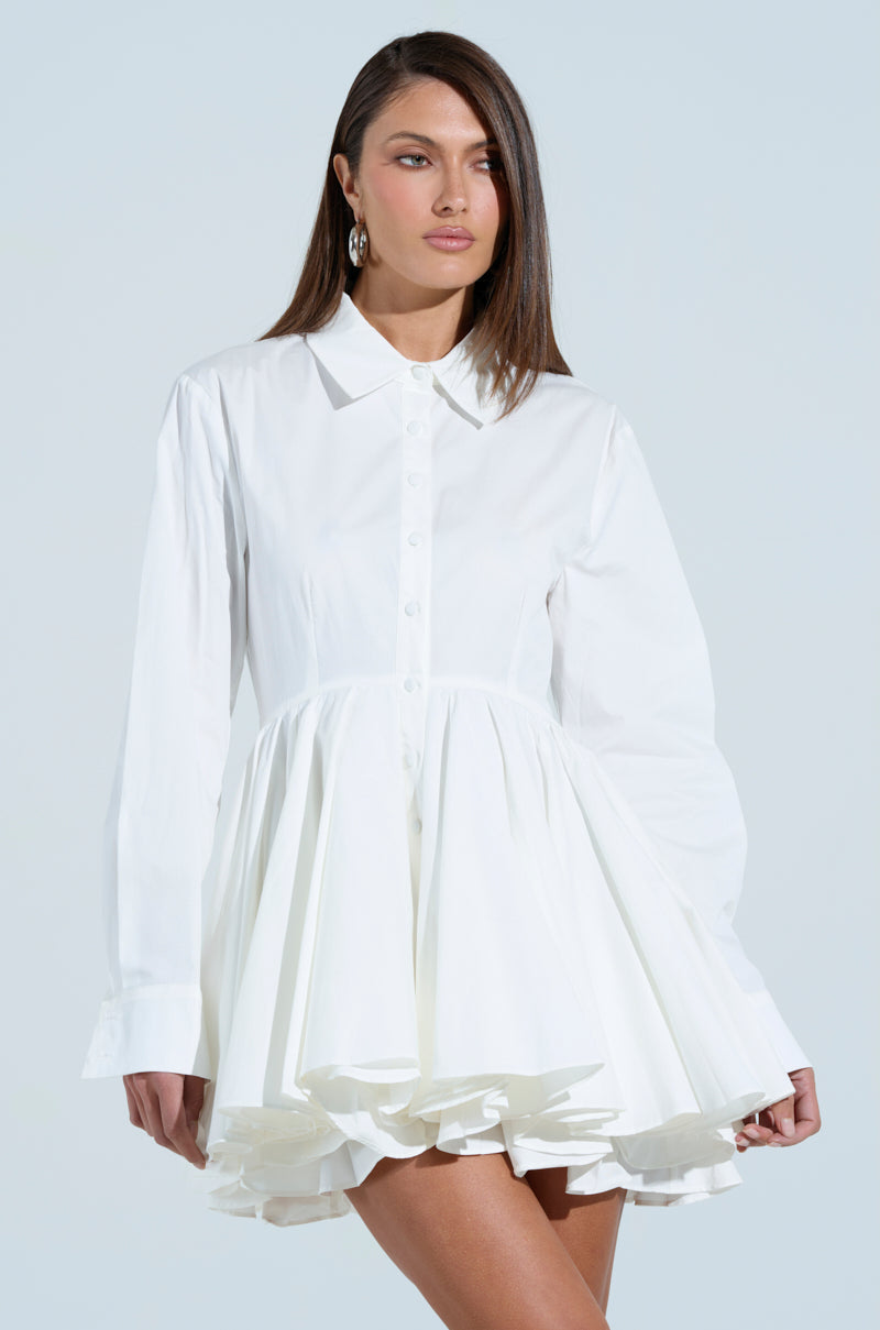 A woman with long brown hair poses in THE IT GIRL POPLIN MINI DRESS, featuring a flared skirt, against a light background. She looks to the side with a neutral expression.