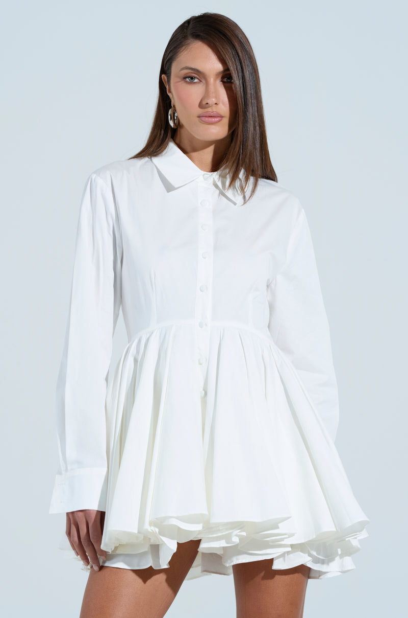 A person with long hair wears THE IT GIRL POPLIN MINI DRESS, showcasing its flared, ruffled skirt against a light background. They gaze directly at the viewer with a neutral expression.