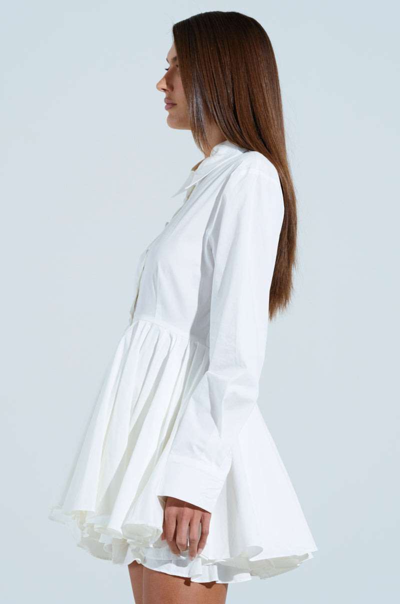 A woman with long brown hair stands in profile against a plain background, wearing THE IT GIRL POPLIN MINI DRESS, a white, long-sleeved skater dress that gracefully flares at the waist.