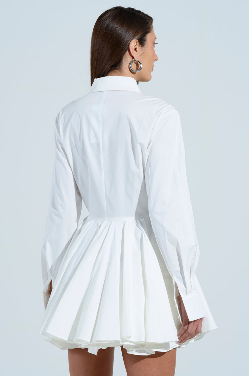 A woman is shown from behind in THE IT GIRL POPLIN MINI DRESS, a white skater dress with a fitted waist and pleated skirt. She has long brown hair, hoop earrings, and stands against a light gray background.