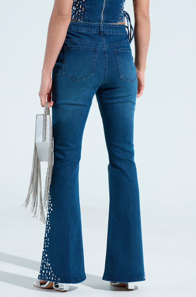 Someone wearing ROCKSTAR DENIM RHINESTONE FLARED PANT and a sleeveless top holds a silver handbag with long fringes. The embellished pants, with designs near the pockets and ankle, exude a rockstar vibe as they pose against a light background.