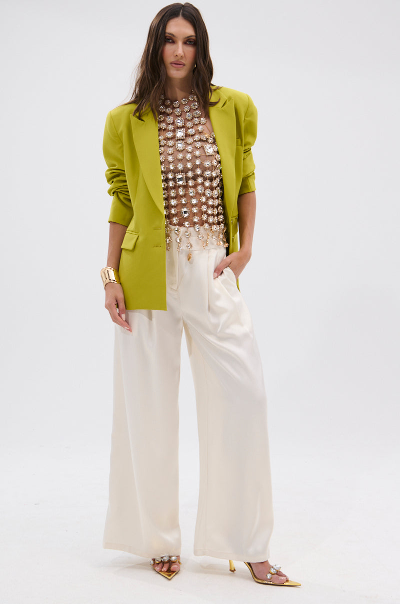 A woman wears the ICY GIRL RHINESTONE EMBELLISHED TOP IN GOLD under a chartreuse blazer, styled with high-waisted white wide-leg trousers, yellow heels, and a chunky gold bracelet. She poses against a plain white background.