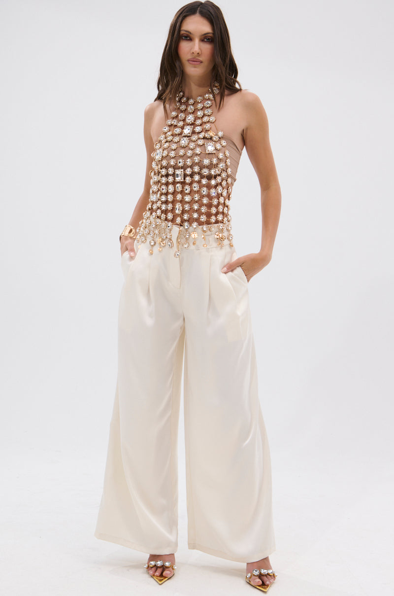 A woman stands against a white background wearing the ICY GIRL RHINESTONE EMBELLISHED TOP IN GOLD and high-waisted, wide-leg white pants. Hands in her pockets, she completes the festival-ready look with strappy gold heels.