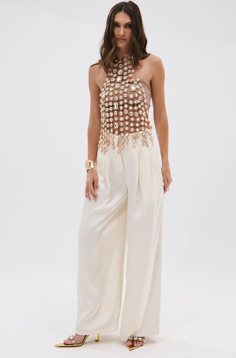 A woman poses in wide-leg white trousers and the ICY GIRL RHINESTONE EMBELLISHED TOP IN GOLD, featuring layered pearl details. She accessorizes with gold heels and a chunky cuff bracelet against a plain white background.