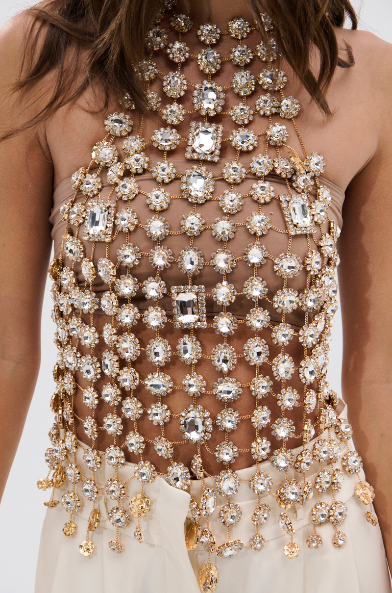 A close-up of a person wearing the ICY GIRL RHINESTONE EMBELLISHED TOP IN GOLD, featuring a grid-like pattern with sparkling crystals and jewels, paired with high-waisted cream pants—perfect for festivals.