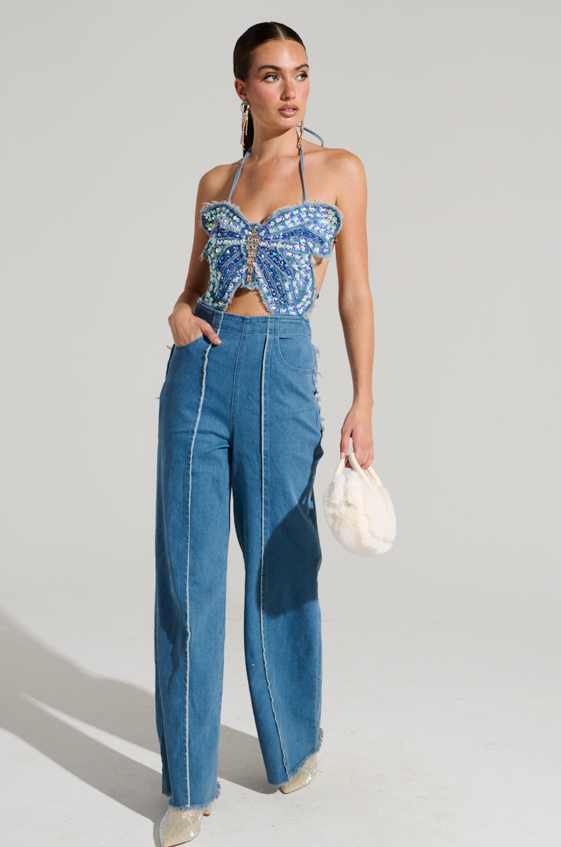 A woman exudes confidence in the SPREAD YOUR WINGS DENIM BUTTERFLY JUMPSUIT, featuring a butterfly-shaped halter top embellished with iridescent sequins. The outfit is paired seamlessly with a small white handbag and light-colored shoes, all set against a plain backdrop. Her hair is styled back, drawing attention to her earrings.