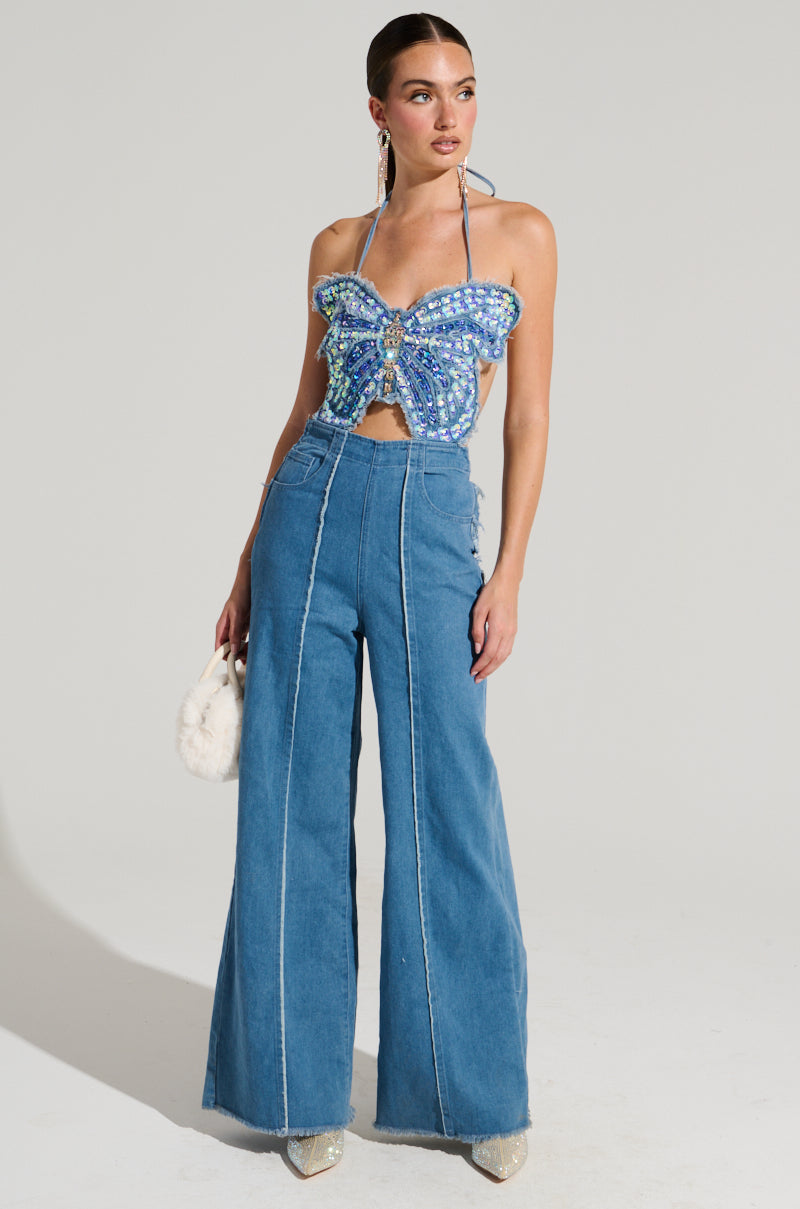 A woman strikes a pose against a simple backdrop, dressed in the SPREAD YOUR WINGS DENIM BUTTERFLY JUMPSUIT. She holds a small white purse and sports large earrings, with her hair neatly pulled back.