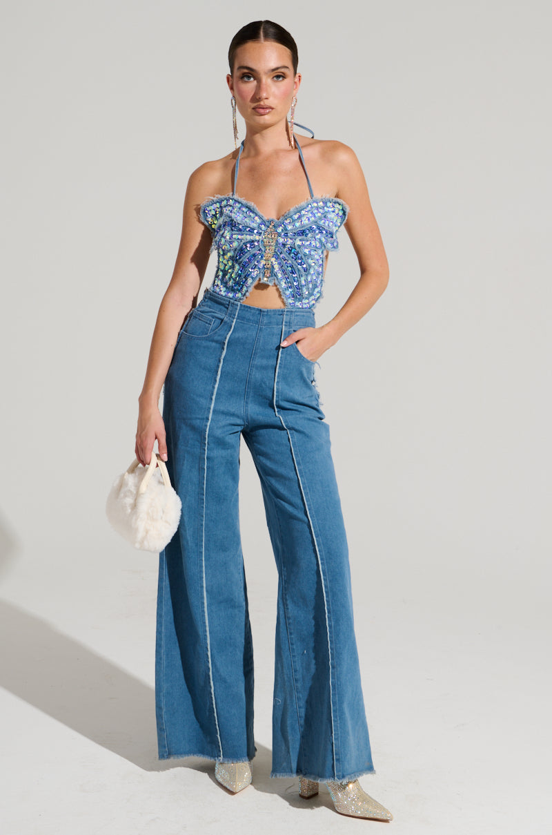 Dressed in the SPREAD YOUR WINGS DENIM BUTTERFLY JUMPSUIT, a woman exudes confidence against a plain backdrop. The jumpsuit features high-waisted denim pants with frayed hems and an iridescent sequin-adorned butterfly-shaped top. Completing her stylish look are light-colored boots, a small white bag, and her hair pulled back effortlessly.