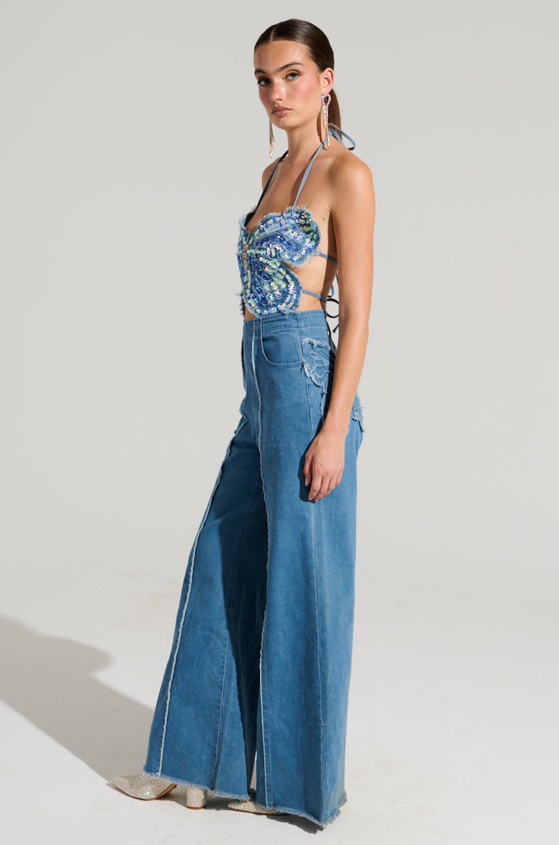 In a creative studio, a person stands confidently in the SPREAD YOUR WINGS DENIM BUTTERFLY JUMPSUIT, featuring vibrant designs and wide-leg jeans. Their long hair is tied back, and they accessorize with iridescent sequin earrings and transparent high-heeled shoes, adding a touch of sparkle to the ensemble.