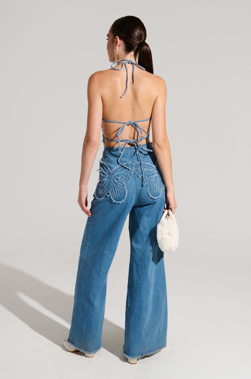A woman wearing the SPREAD YOUR WINGS DENIM BUTTERFLY JUMPSUIT, featuring a halter top with lace-up back details and wide-leg jeans adorned with iridescent sequin butterfly designs, stands facing away holding a small white bag against a plain background. Her hair is styled in a sleek ponytail.