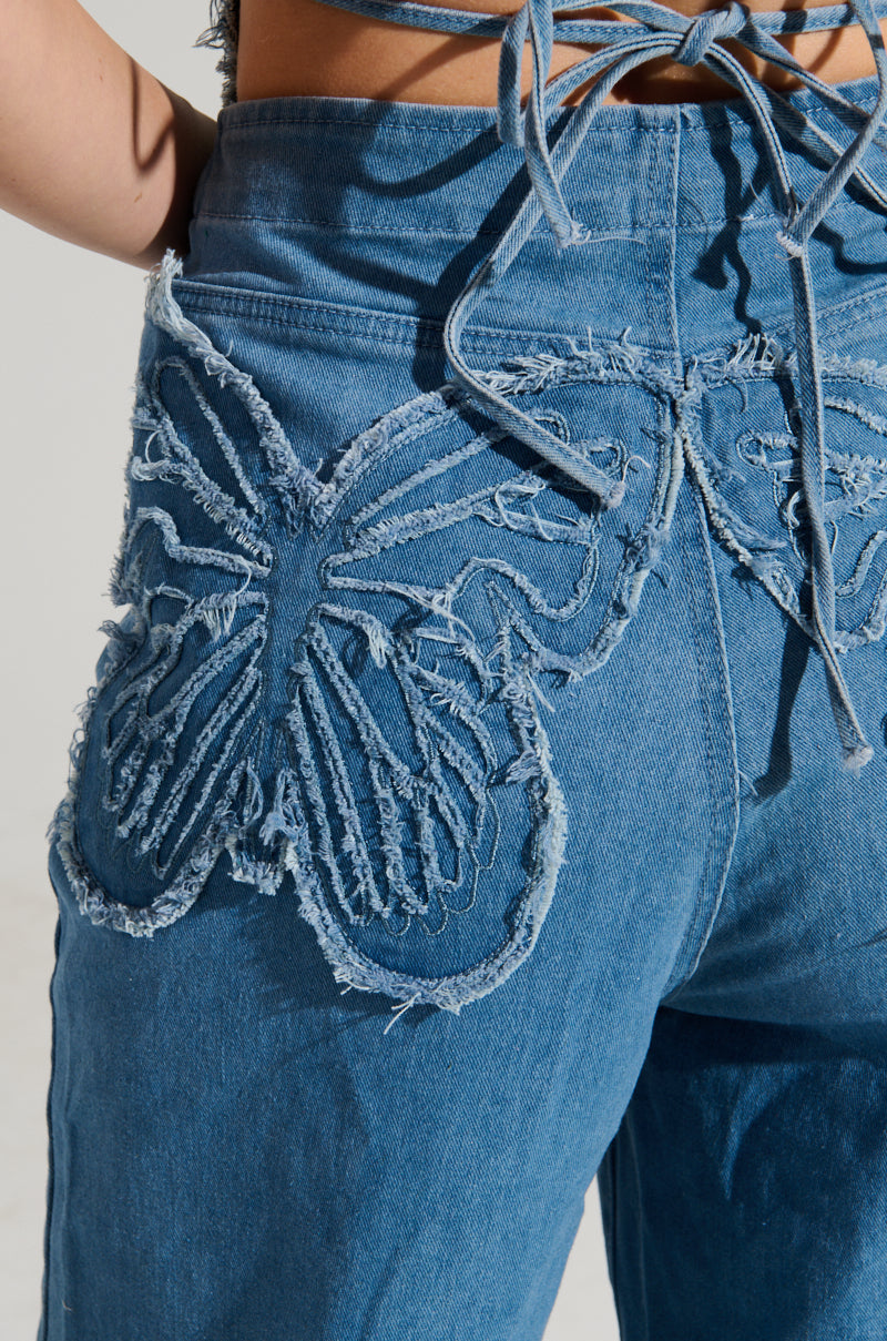Close-up of the SPREAD YOUR WINGS DENIM BUTTERFLY JUMPSUIT, highlighting a large, frayed butterfly design on the back pocket. Iridescent sequins add a touch of sparkle, while the crisscross tie detail on the waistband completes this artistic and textured look.