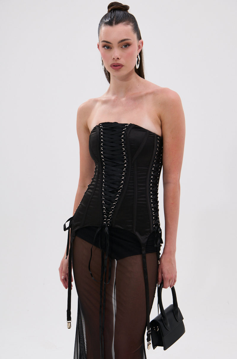 A woman with long, dark hair in a high ponytail wears the LACE ME UP CORSET— a black satin corset top with lace-up details—paired with a sheer black skirt, large hoop earrings, and holds a small black handbag against a white background.