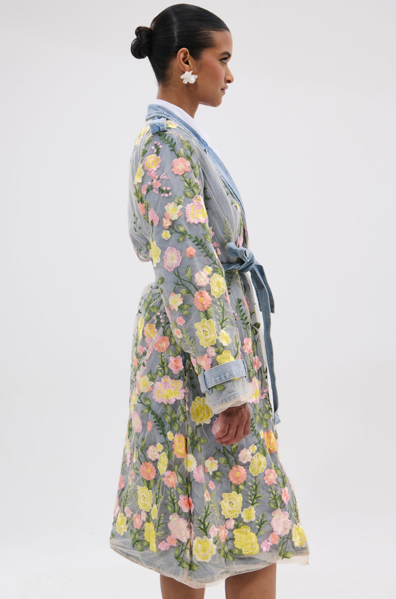 A woman stands in profile wearing the LIFE IN COLOR FLORAL DENIM TRENCH, a sheer, knee-length coat with pink, yellow, and green floral embroidery. The cinched silhouette flatters her figure against a plain light backdrop.