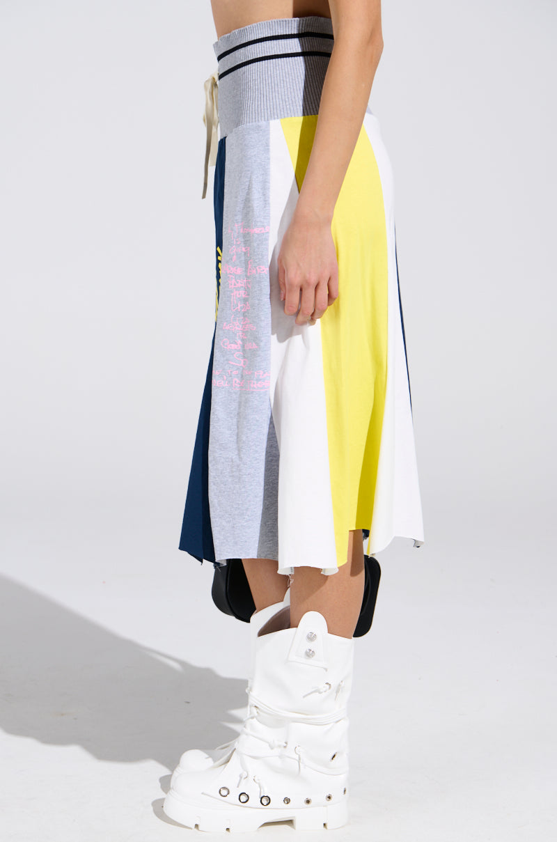 A person wearing the REBUILT MIDI SKIRT, a sporty-chic knee-length piece with gray, white, yellow, and navy color-block panels, paired with tall white boots. The side photo shows only the torso down.