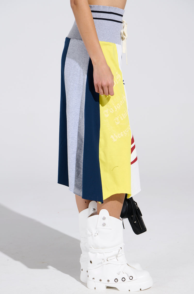 A person stands sideways wearing the REBUILT MIDI SKIRT, a sporty-chic color-block design with yellow, gray, navy, and white panels, paired with tall white boots featuring silver buckles and a ribbed waistband.