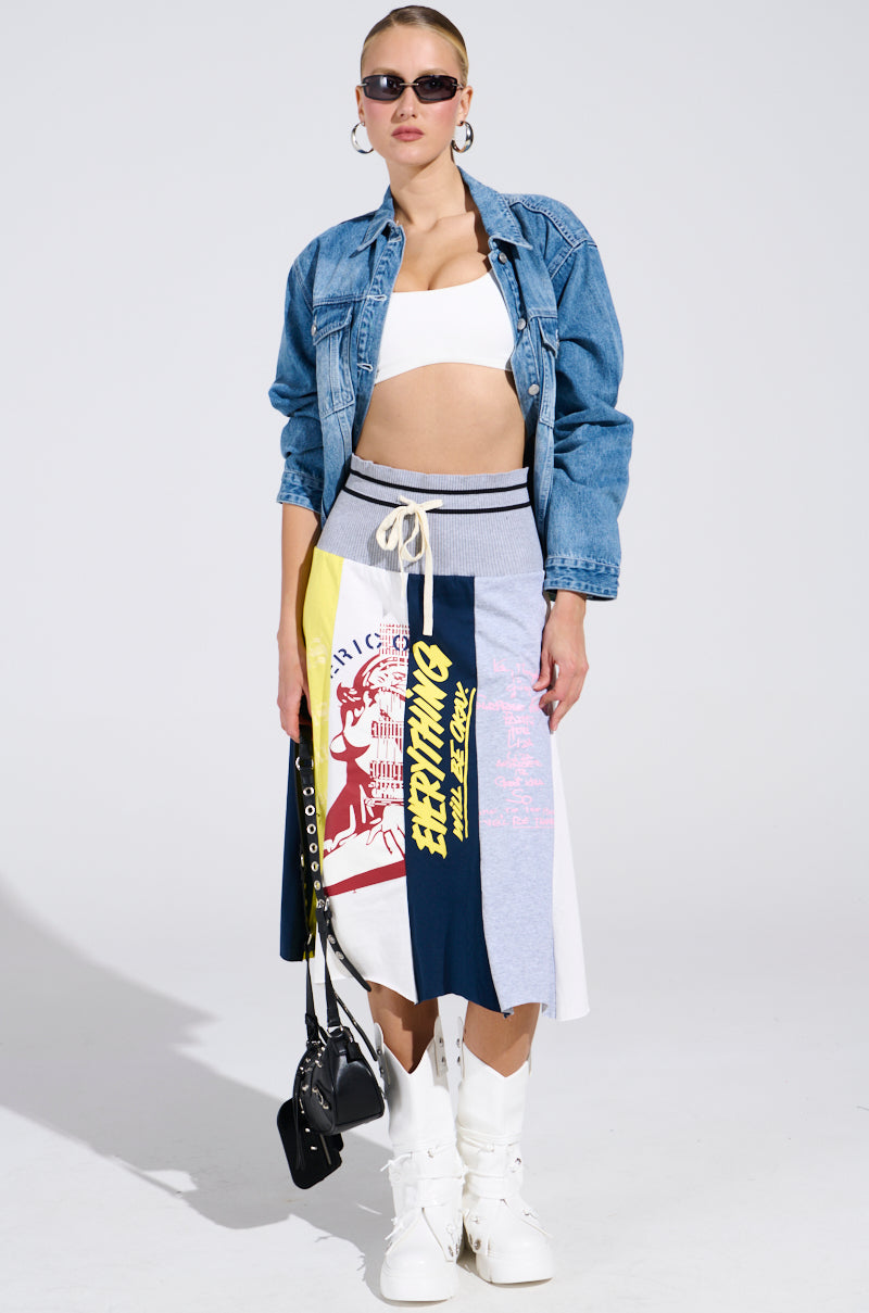 A woman poses confidently against a plain background, wearing sunglasses, a denim jacket over a white crop top, the REBUILT MIDI SKIRT with text detail, white knee-high boots, and holding a black handbag.