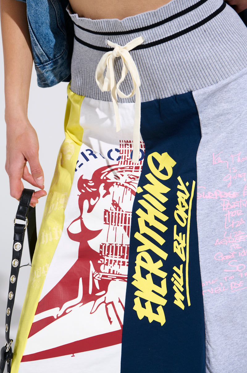 A close-up of a person wearing the REBUILT MIDI SKIRT, featuring colorful patchwork, bold yellow "EVERYTHING WILL BE OKAY" text, abstract graphics, and styled with a denim jacket for sporty-chic flair.