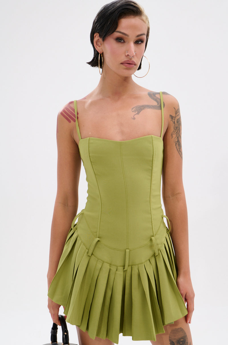 A woman with short black and blonde hair wears the DANCING THROUGH LIFE MINI DRESS IN OLIVE, featuring thin straps and a corset-style bodice. She accessorizes with tattoos, large hoop earrings, and a small black handbag against a white background.