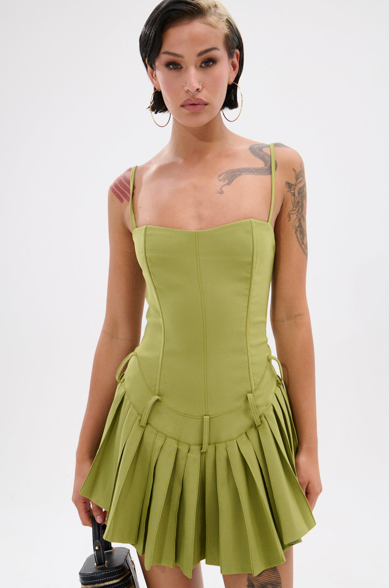 A woman with short dark hair and hoop earrings wears the DANCING THROUGH LIFE MINI DRESS IN OLIVE, featuring thin straps and a corset-style bodice, paired with a small black handbag, standing against a plain white background.