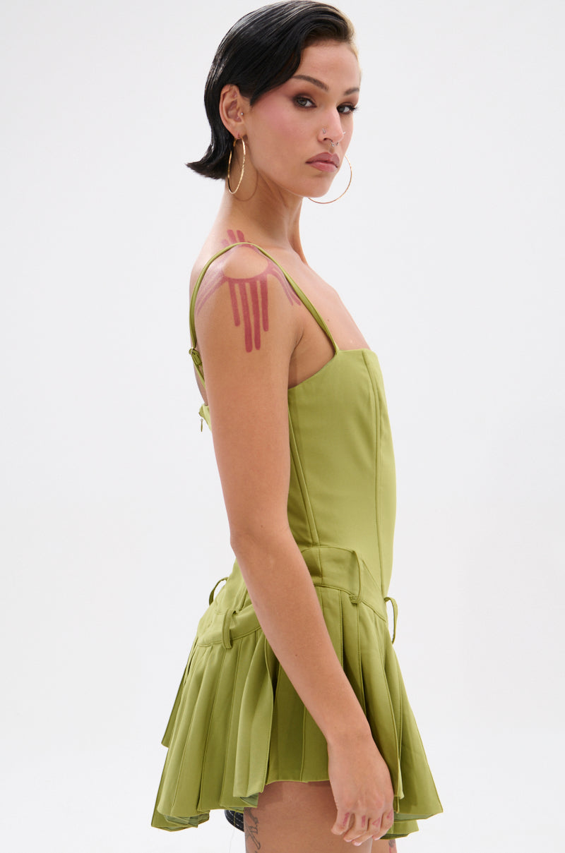 A person with short dark hair and hoop earrings wears the DANCING THROUGH LIFE MINI DRESS IN OLIVE, featuring a corset-style bodice. Pink paint-like marks are on their right shoulder as they look over their shoulder against a white background.