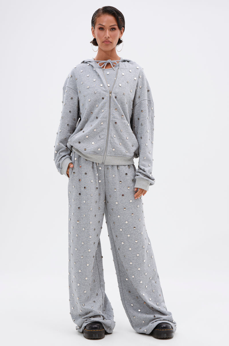 A woman faces forward in the POPPING OFF ZIP UP SWEATSHIRT—a loose hooded piece with stud embellishments—paired with matching wide-leg pants. She has slicked-back hair, black shoes, and stands against a plain white background.