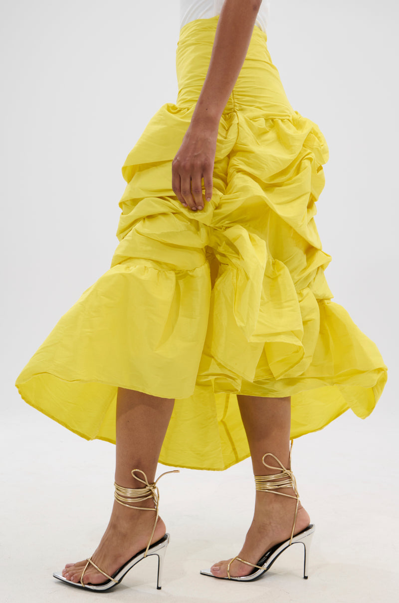 A model walks forward against a plain white background, wearing the SUNSHINE MIDI SKIRT in bright yellow with ruffles and asymmetrical draping, paired with strappy high-heeled sandals.