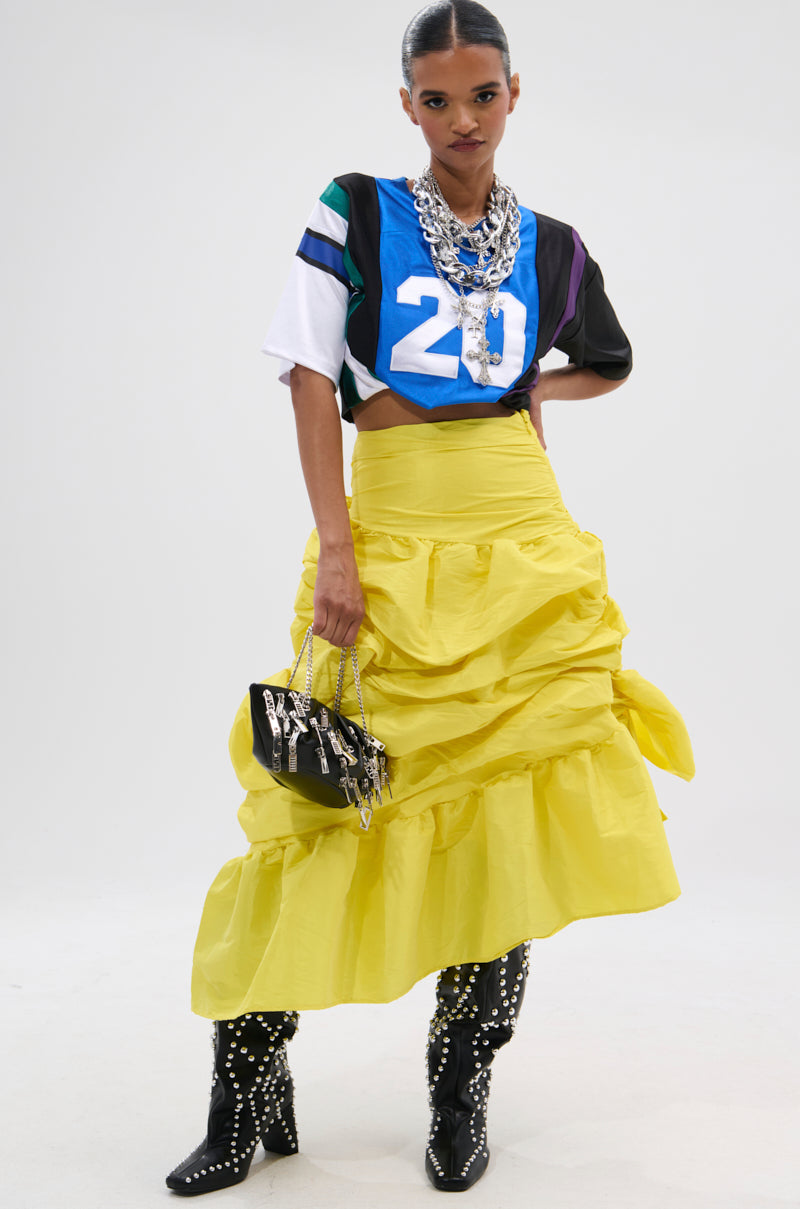 A woman poses wearing the SUNSHINE MIDI SKIRT in bright yellow, paired with a blue and white number 20 sports jersey, black studded boots, layered silver necklaces, and carrying a small black handbag.