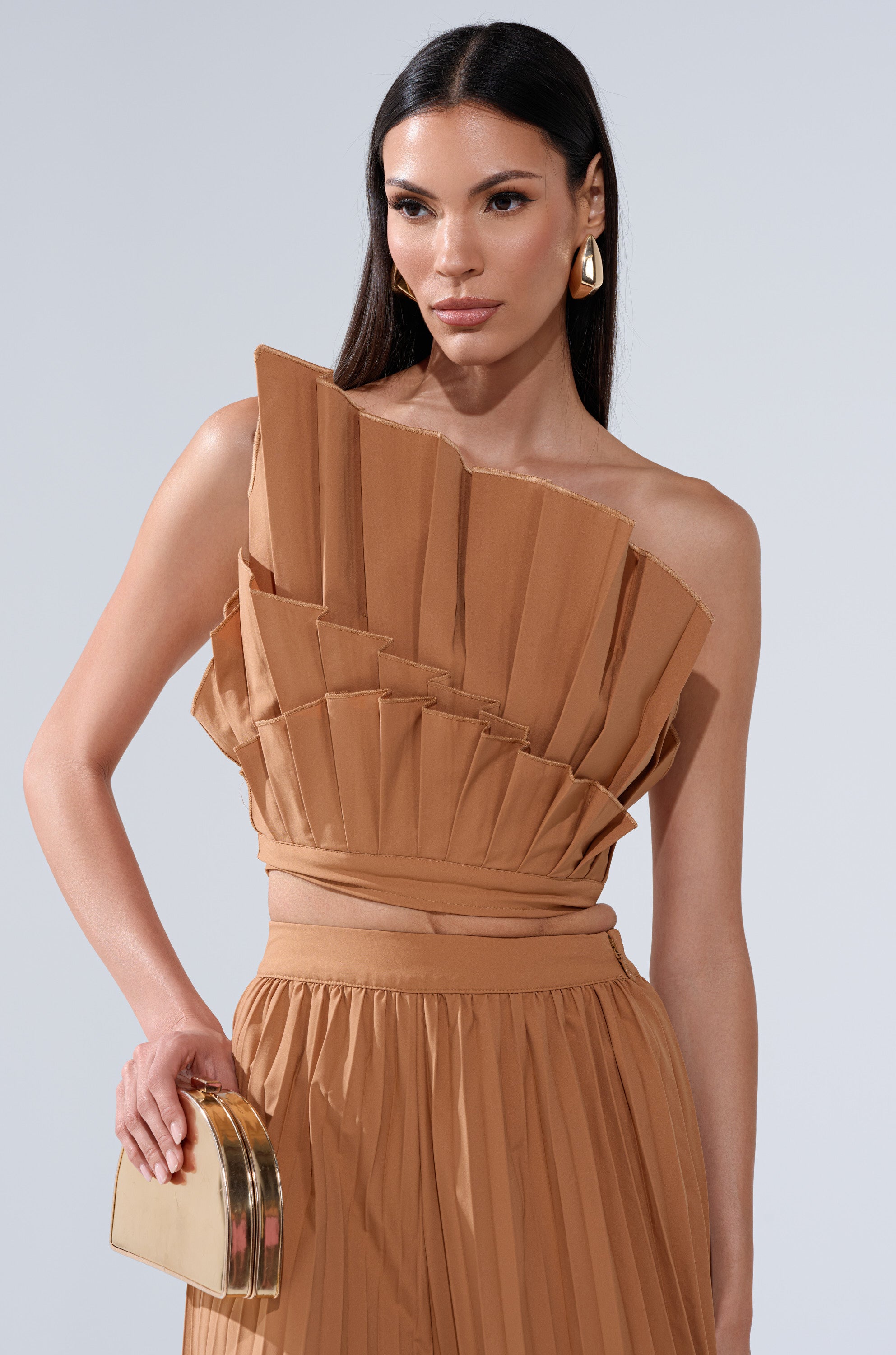 A woman with long dark hair models the STAR OF THE NIGHT PLEATED BLOUSE—an asymmetrical, one-shoulder tan top—styled with gold hoop earrings and a gold clutch against a plain light background.