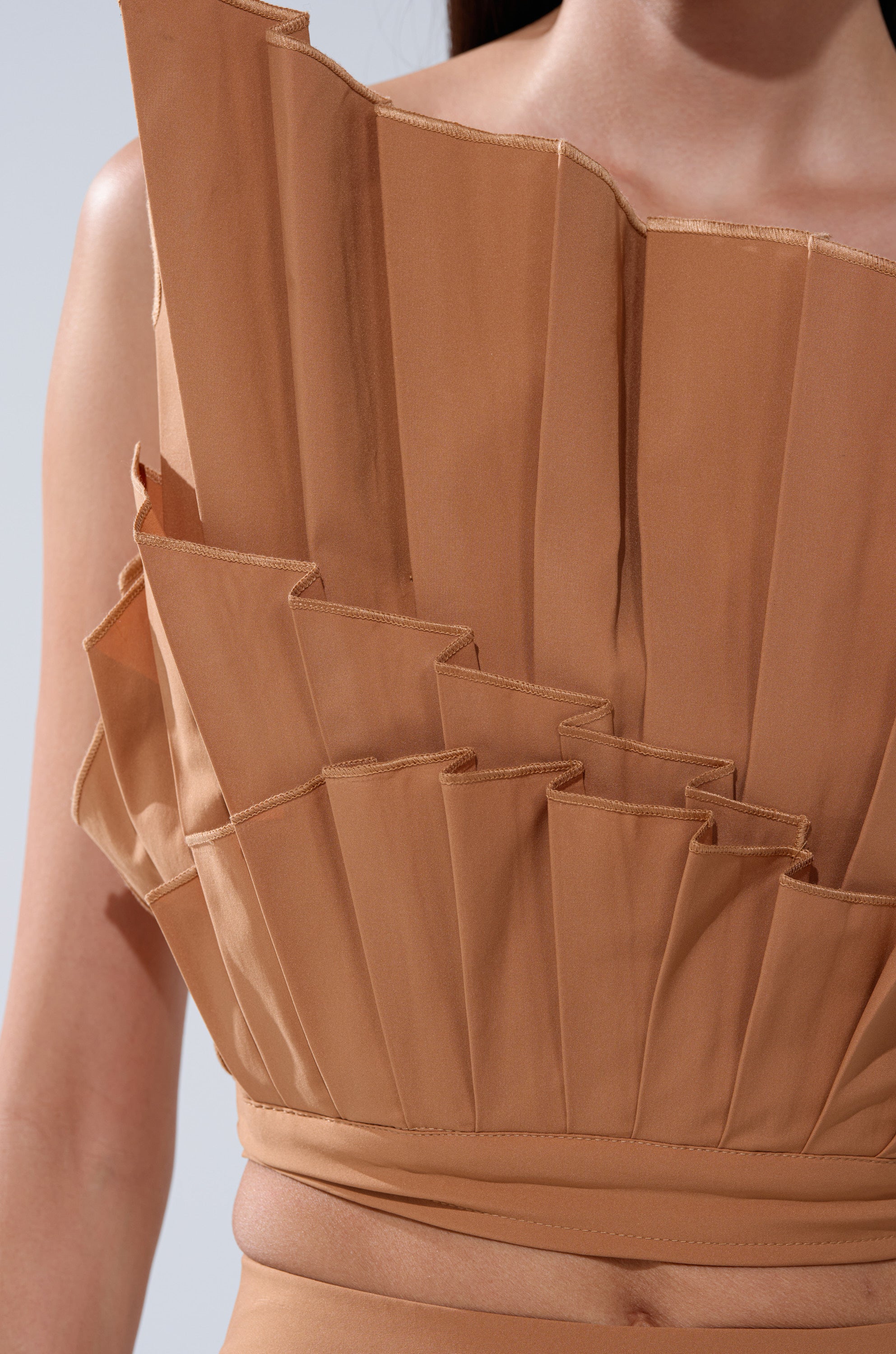 A close-up shows someone wearing the STAR OF THE NIGHT PLEATED BLOUSE, featuring tan fabric with layered, geometric pleats and sharp, angular edges. This architectural design offers a modern twist to evening wear. The person's head is not visible.