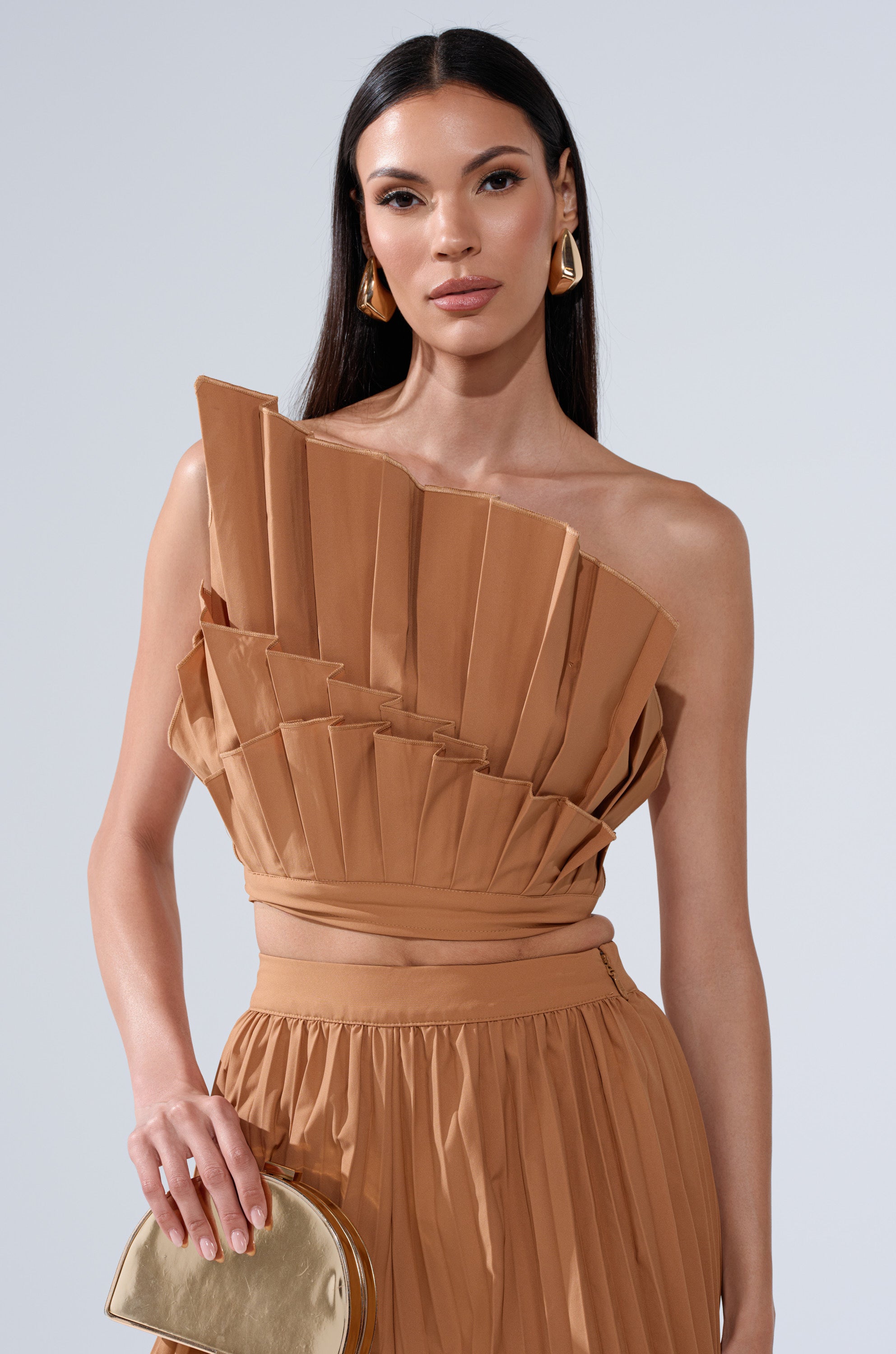 A woman with long dark hair wears the tan STAR OF THE NIGHT PLEATED BLOUSE and matching skirt, styled with gold earrings and a small gold clutch. She poses against a plain, light gray background.