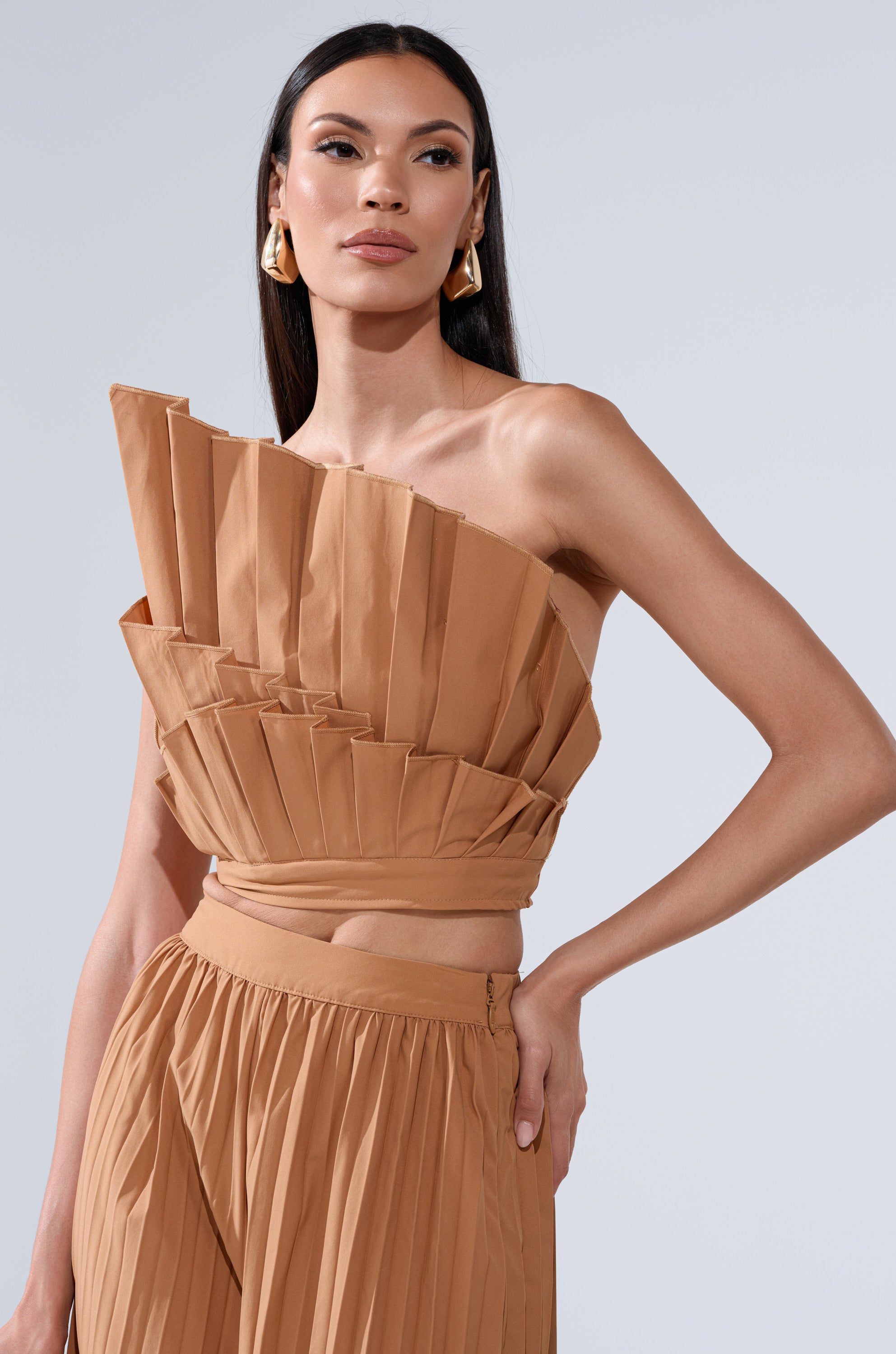 A woman wearing the STAR OF THE NIGHT PLEATED BLOUSE and a matching pleated skirt poses confidently against a light gray background, her long dark hair styled straight and accessorized with gold earrings.