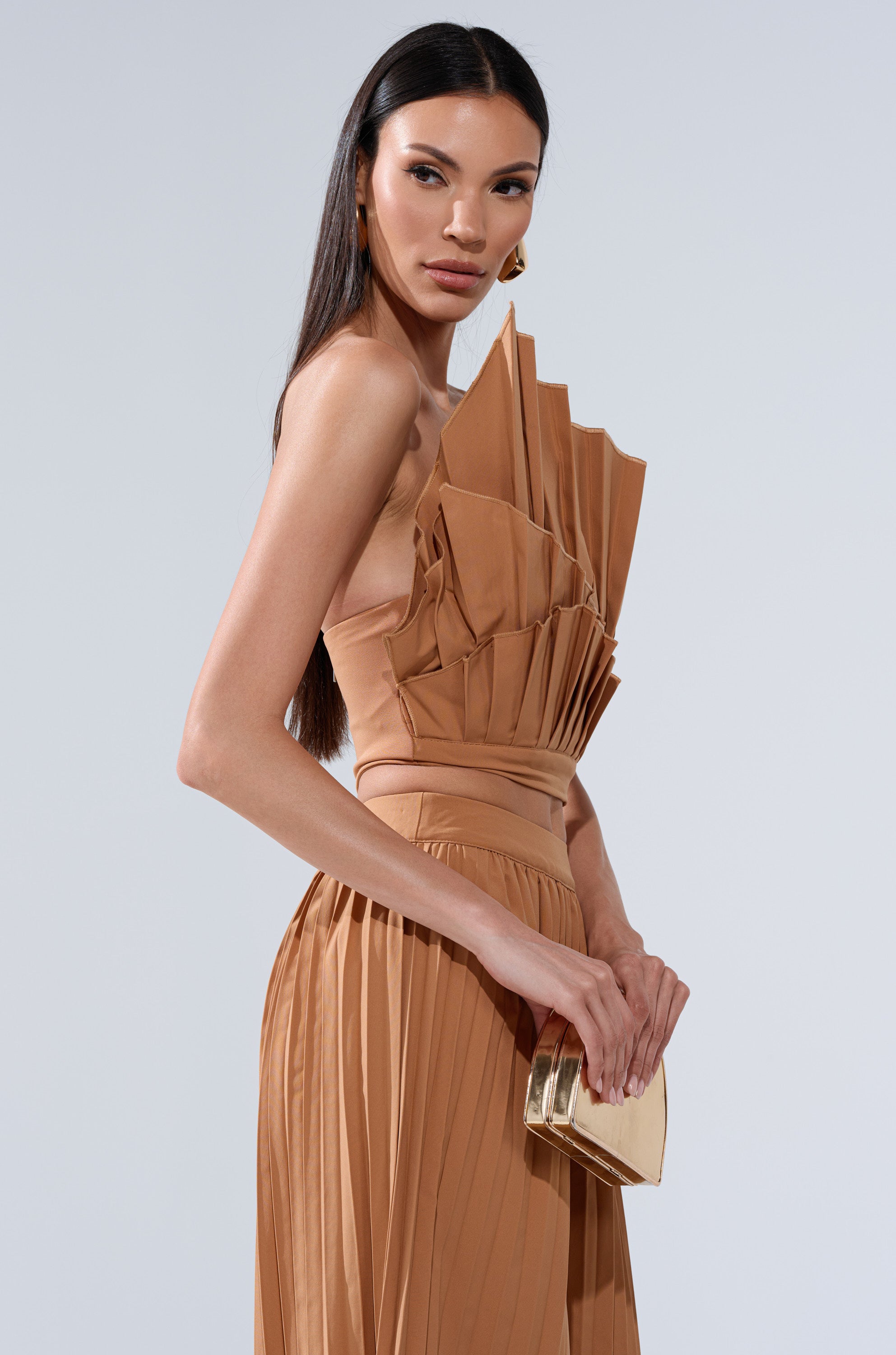 A woman in the STAR OF THE NIGHT PLEATED BLOUSE, featuring a one-shoulder, bronze-brown starburst-inspired design, holds a metallic gold clutch. She has long straight dark hair and poses against a plain light background.
