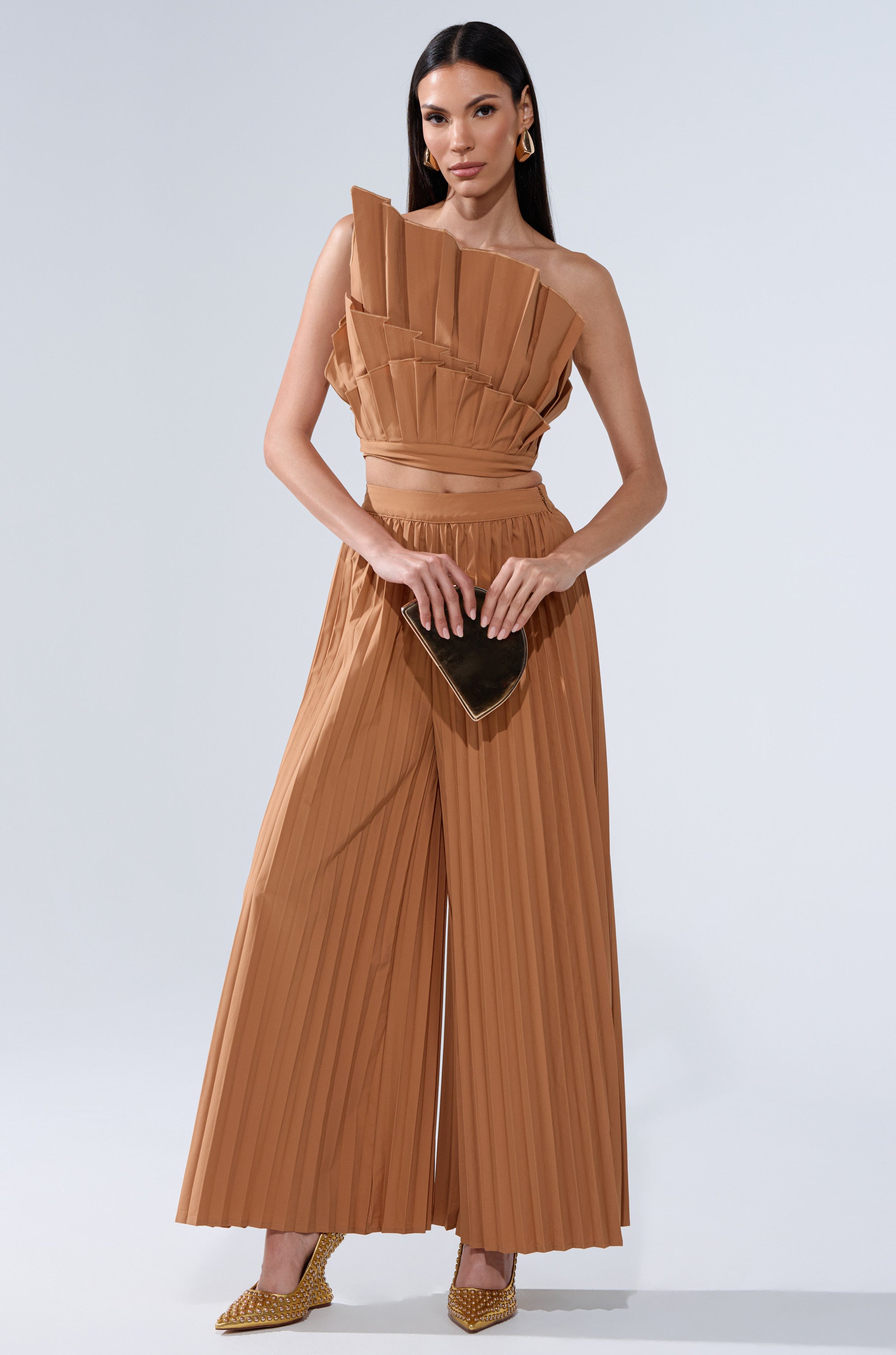 A woman stands confidently in the STAR OF THE NIGHT PLEATED BLOUSE, paired with pointed tan heels and gold earrings. She holds a small black clutch, her long straight hair framing her look against a plain light background.