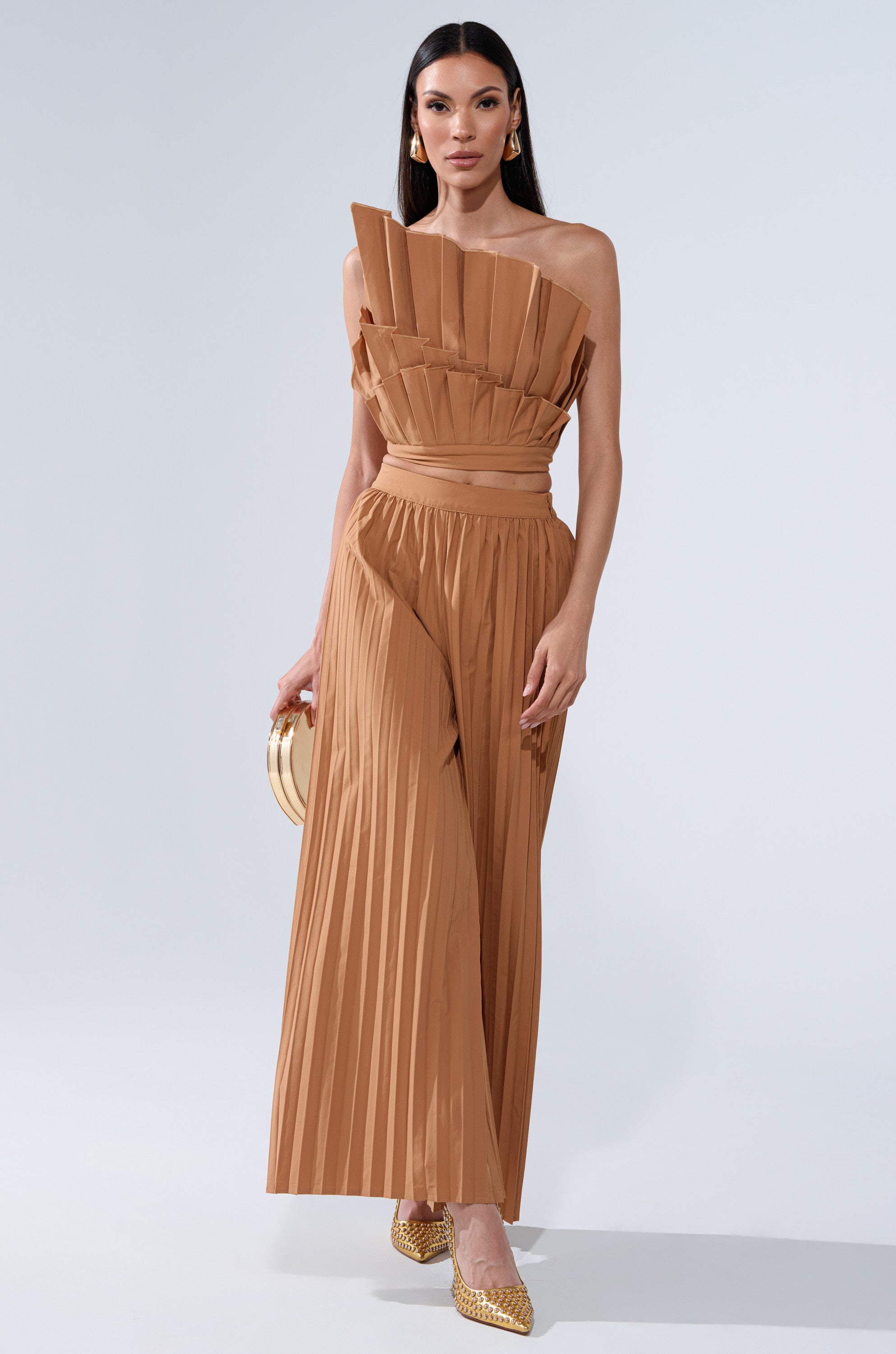 A woman models the STAR OF THE NIGHT PLEATED BLOUSE—a camel-colored, starburst one-shoulder top paired with matching wide-leg pants. She accessorizes with a small round bag and gold pointed heels, standing against a plain backdrop.