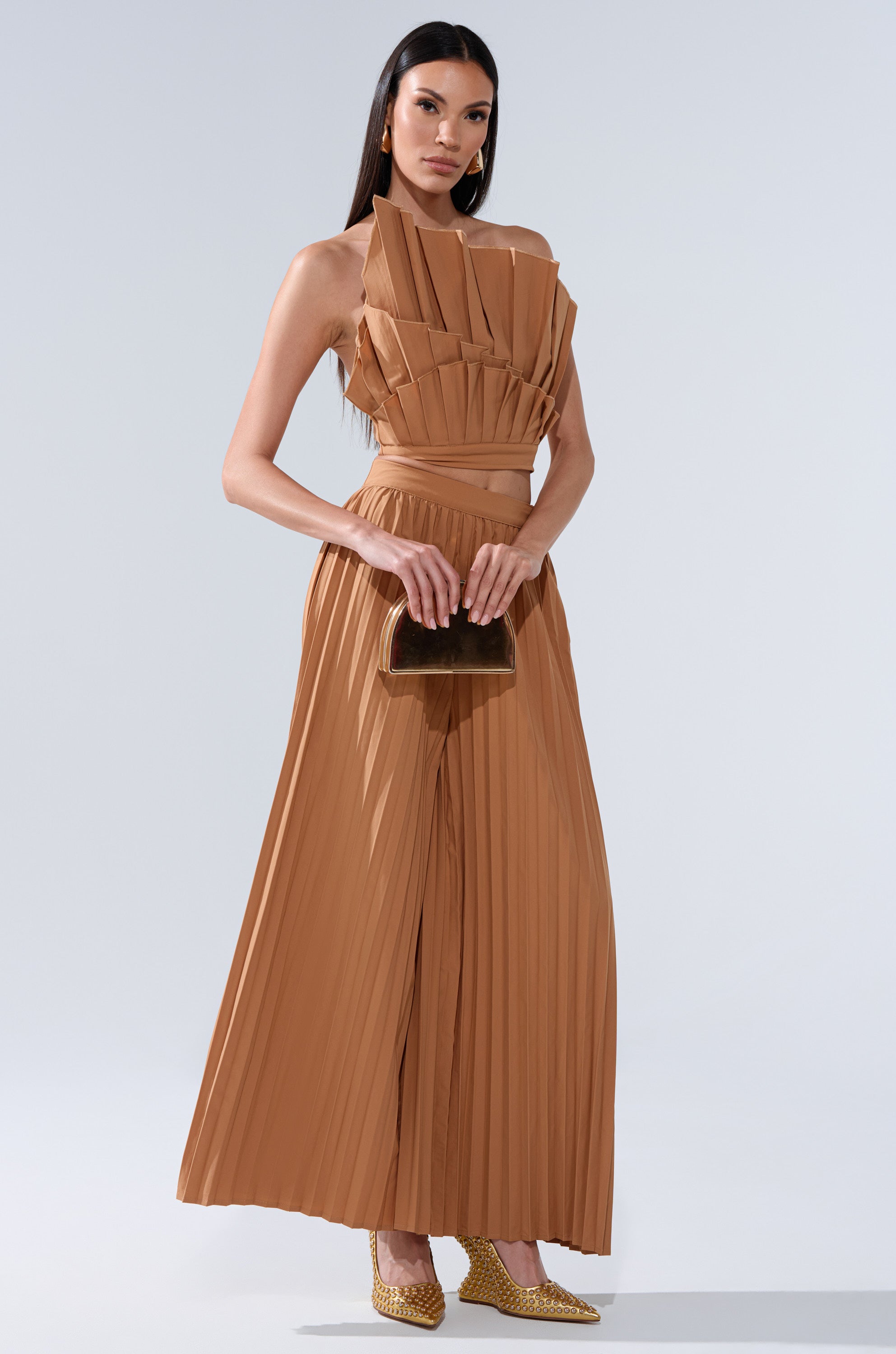 A woman wears the STAR OF THE NIGHT PLEATED BLOUSE, paired with a small brown clutch and pointed gold shoes. Her hair is straight, she has gold earrings, and her look is polished and elegant.